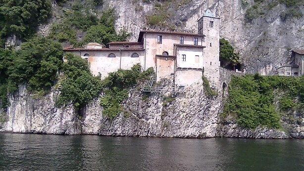 Un edificio de piedra con múltiples ventanas y una torre de campanas, ubicado en un acantilado con vistas a un cuerpo de agua.