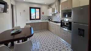 A modern kitchen with a gas stove, oven, microwave, and stainless steel refrigerator. There is a dining table with chairs and a window with a view of buildings. - Archimedes Lounge in Cefalù (Cefalù)