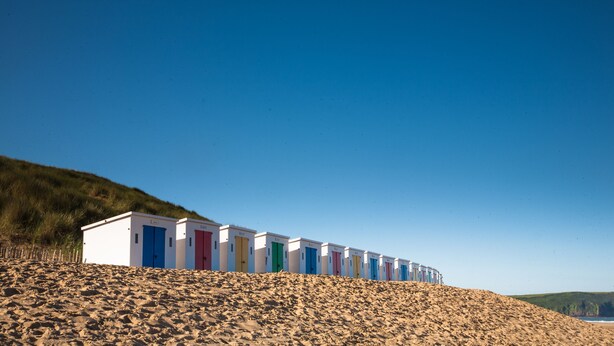 Eine Reihe von Strandhütten mit unterschiedlichfarbigen Türen, die an einem sandigen Hang unter blauem Himmel angeordnet sind.