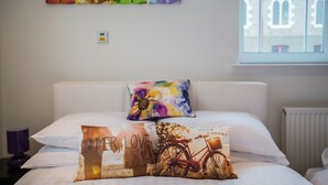 Ein Bett mit weißer Bettwäsche, einem farbenfrohen Kissen und einem Kunstwerk darüber.