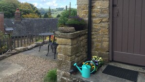 Un edificio de piedra con una puerta marrón, un jardín con flores, una mesa de metal y un regador.