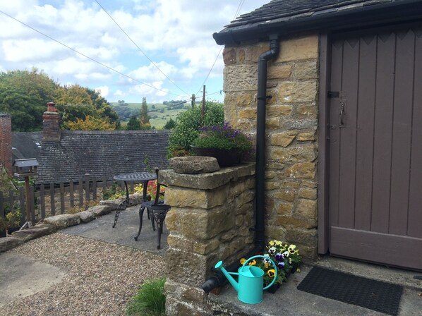A stone building with a brown door, a garden with flowers, a metal table, and a watering can.
