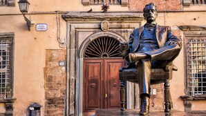 Une statue représentant un homme assis, vêtu d’un costume, est placée devant un bâtiment doté d’une porte en bois et d’une enseigne indiquant « Piazza Cittadella ».