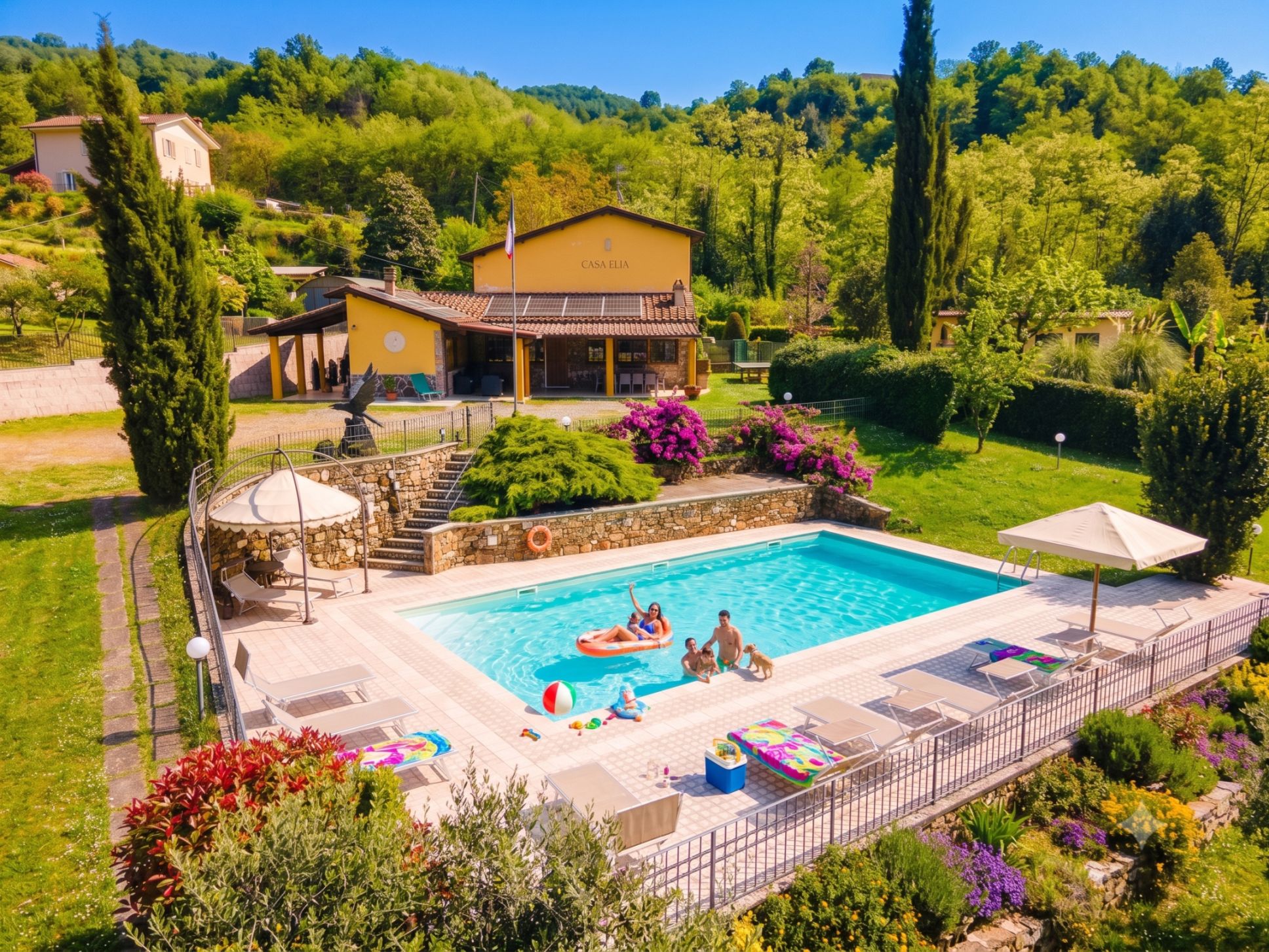 A swimming pool with lounge chairs and a covered seating area, surrounded by well-maintained gardens and a house named 'CASA ELIA'.
