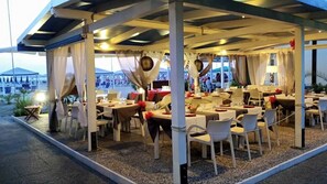 An outdoor restaurant with tables and chairs under a covered area, with a view of the beach in the background.