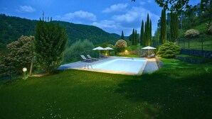 Une piscine entourée de chaises longues et de parasols, bordée d’une pelouse bien entretenue et d’arbres, avec des collines en toile de fond.