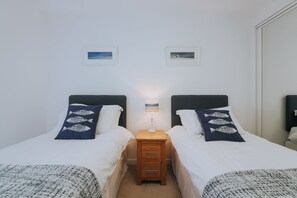 Habitación doble con dos camas, cada una con colcha blanca y almohada estampada con motivos de peces. Entre las camas, una mesita de noche de madera con una lámpara.