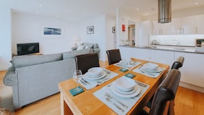 A modern living room with a grey sofa, a dining table set for four, and a kitchen in the background.