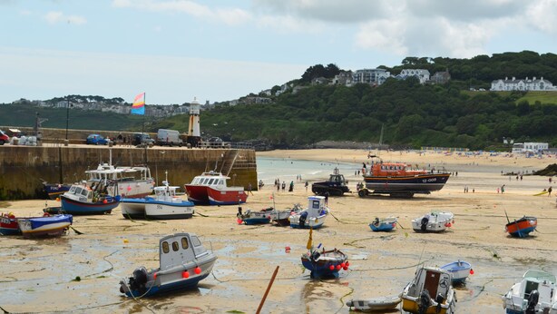 A coastal scene with boats docked at a pier, a lighthouse in the background, and a beach with people.
