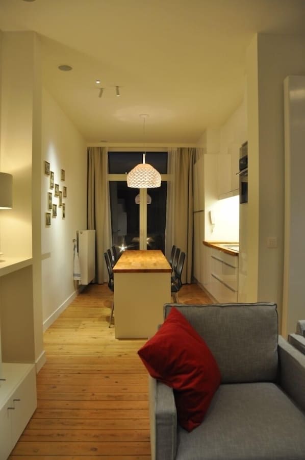 Un espace de vie moderne et compact comprenant un coin repas, une kitchenette et un canapé agrémenté d’un coussin rouge.