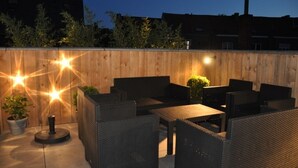 Een terras buiten met rieten meubilair en potplanten, omringd door een houten muur en een stadsgezicht op de achtergrond.