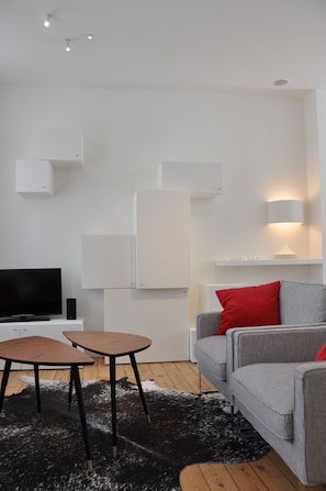 Un salon moderne avec un téléviseur à écran plat, deux tables en bois, un canapé gris avec des coussins rouges et un tapis noir et blanc.
