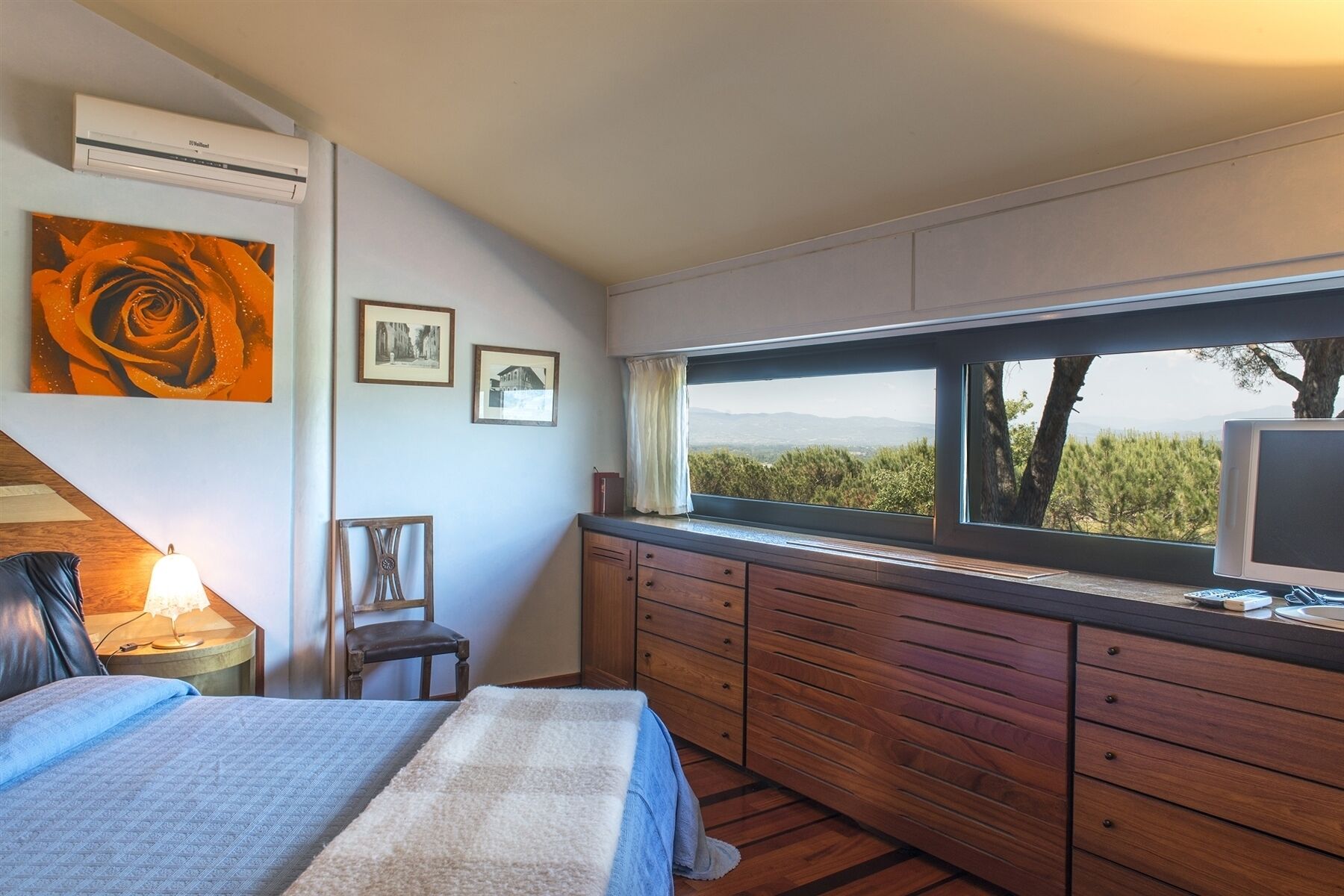 A bedroom with a large window, a bed, a dresser, and artwork on the wall.