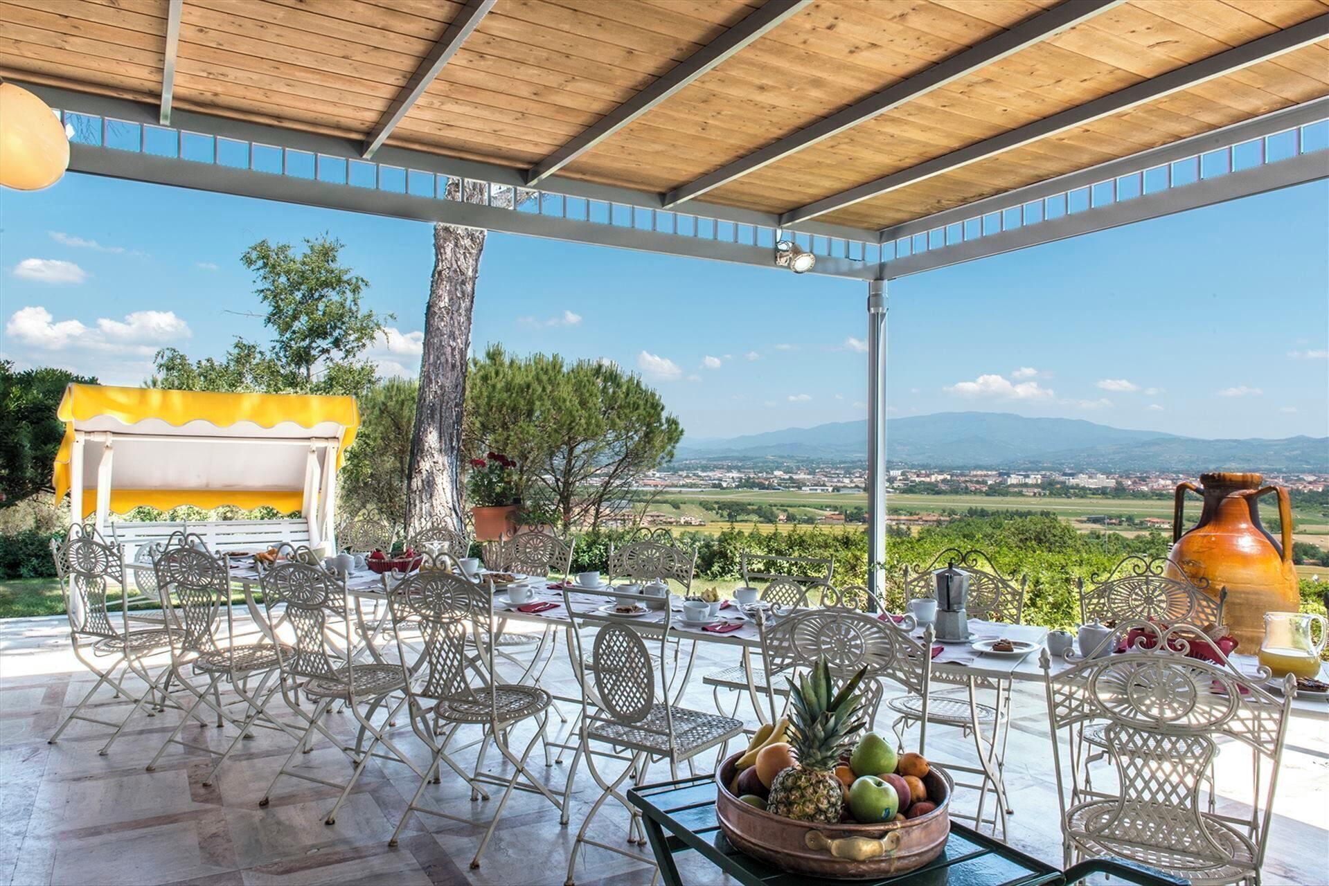 An outdoor patio with metal chairs and tables, a yellow awning, and a view of the countryside.