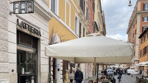 Tavoli all'aperto con sedie in metallo bianco e un ombrellone grande, una strada acciottolata e edifici dall'architettura classica.