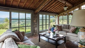 A spacious living room with a stone wall, large windows offering a view of the countryside, a sofa, a coffee table, and a decorative lamp. - Exceptionally pretty cottage with pool, views, in walking distance from Panzano (Greve in Chianti)