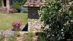A stone building with a tiled roof, surrounded by a garden with flowering plants and a clear blue sky. - Exceptionally pretty cottage with pool, views, in walking distance from Panzano (Greve in Chianti)