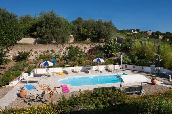 A swimming pool with lounge chairs and umbrellas, surrounded by greenery and a hillside.