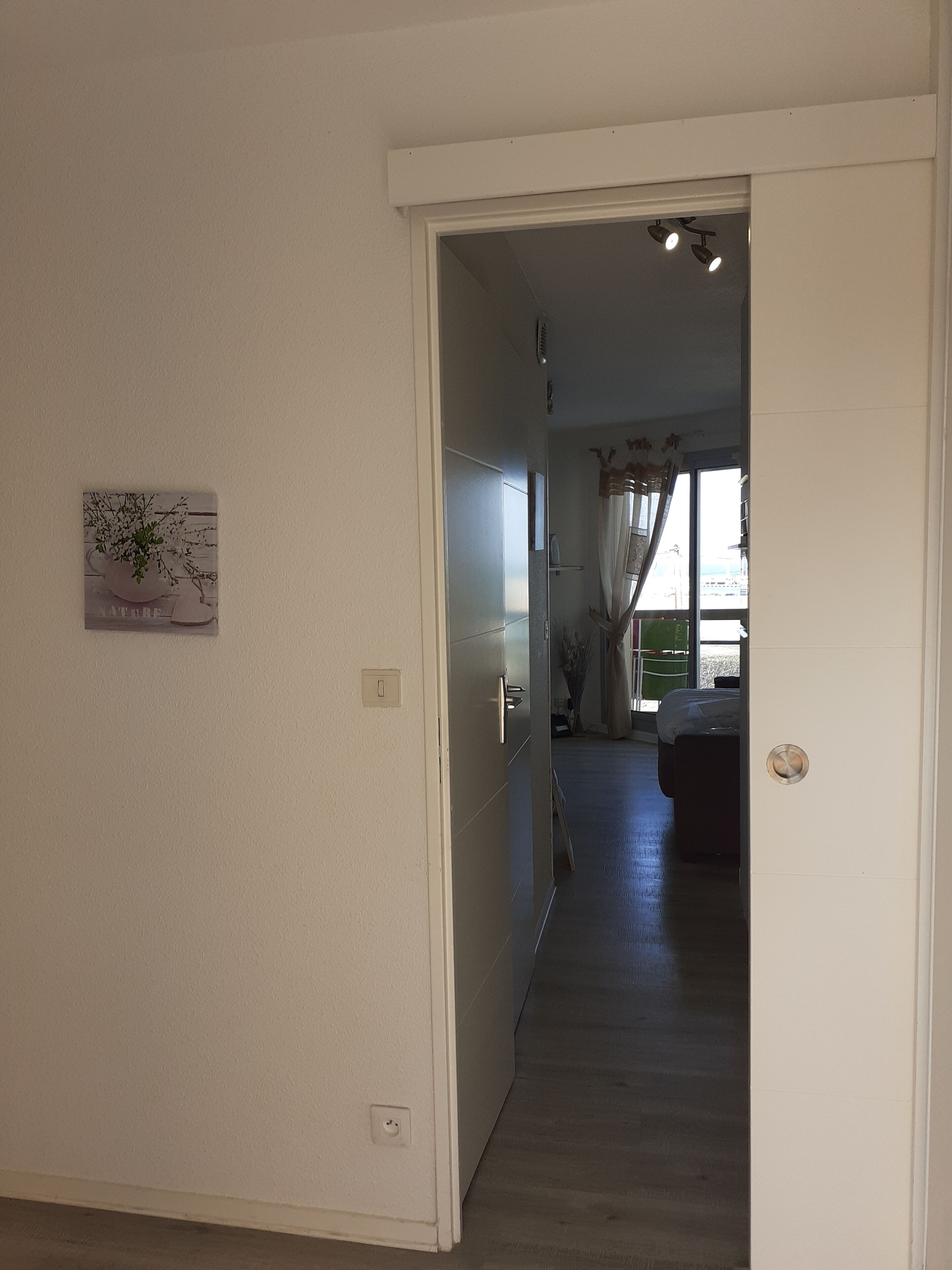 Un pasillo que conduce a un dormitorio con una cama, una ventana con cortinas y una lámpara fijada a la pared.