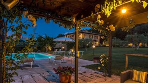 Un espace extérieur aménagé avec une terrasse, une piscine, des chaises longues et une pergola illuminée de guirlandes lumineuses.