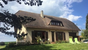 A two-story house with a sloped roof, a front porch, and a lawn with two green lawn chairs. - Norman house with garden near the landing beaches  (Ver-Sur-Mer)