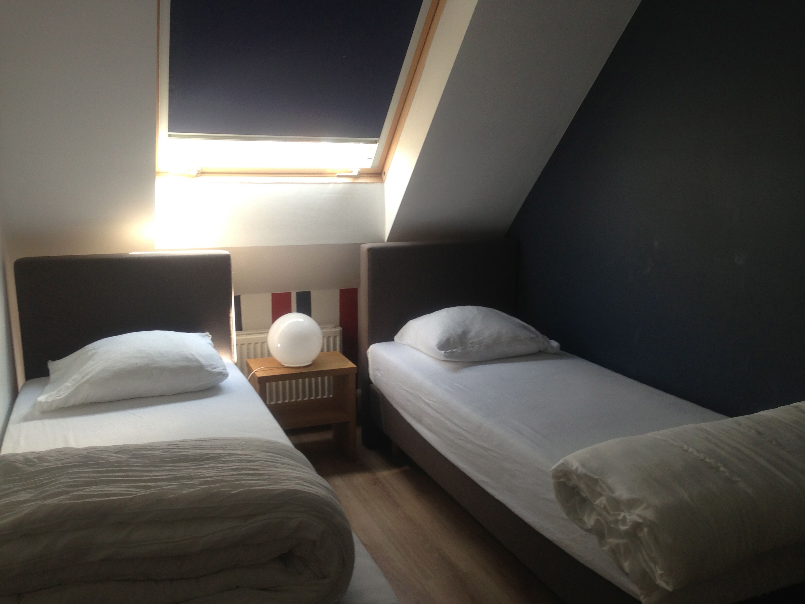 Quarto pequeno e pouco iluminado, com duas camas de solteiro, uma mesinha com abajur e um radiador.