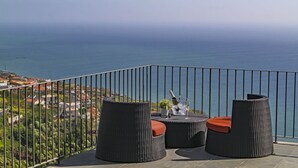 A balcony with wicker furniture and a bottle of champagne, overlooking a coastal view. - Villa Clementina | Cliffs & Ocean | Heated Pool 26ºC (Prazeres)