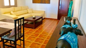 Ein Wohnzimmer mit einer Couch, einem Couchtisch, einem Esstisch und einem Stuhl. An der Wand hängt ein Gemälde, im Hintergrund ist eine Tür zu sehen.