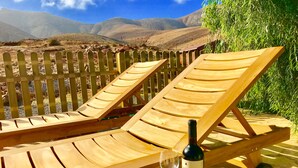 Zwei Holz-Loungestühle mit einer Weinflasche und einem Glas auf einem Holztisch, vor einer Berglandschaft.