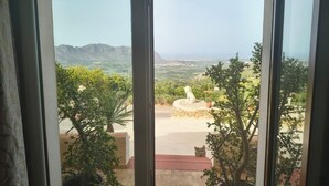 Vista desde una ventana con una estatua de piedra y plantas en macetas en primer plano, y un paisaje montañoso al fondo.