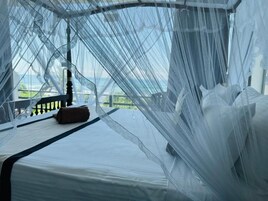 Una cama con sábana blanca, almohada marrón, mosquitero y vistas a la vegetación.