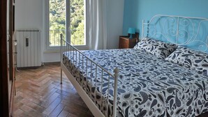 Een slaapkamer met een bed, een nachtkastje, een raam met uitzicht op groen en een radiator.