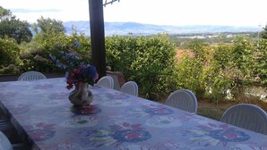 Een terras met een tafel gedekt voor vier personen, omringd door stoelen. Op de tafel staat een vaas met bloemen. Achtergrond: een landschap met bomen en bergen.