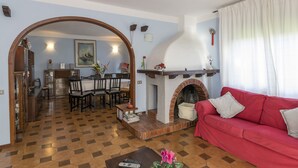 Een woonkamer met een rode bank, een open haard, een eethoek met stoelen en een tafel, en een gang die naar een andere kamer leidt.