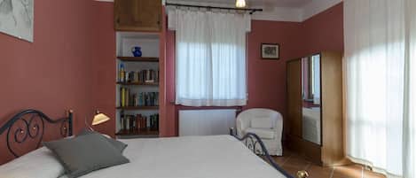 A bedroom with a bed, a bookshelf, a chair, a radiator, and a window with curtains.