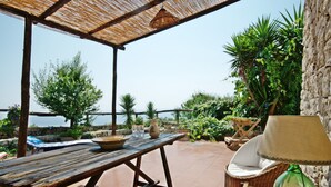 Een houten tafel met een kom en karaf, een rieten stoel met kussen, en een lamp op een terras met uitzicht op zee.