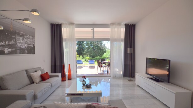 A modern living room with a sofa, coffee table, and TV. There is a glass door leading to an outdoor patio with chairs and a table. - House, 3 terraces, directly on golf course, swimming pool, tennis. Close to beach (Nueva Andalucia,)