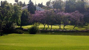 A golf course with a pond, trees, and a building in the background. - House, 3 terraces, directly on golf course, swimming pool, tennis. Close to beach (Nueva Andalucia,)
