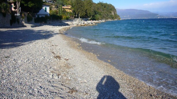 Une plage de galets avec une eau claire, l’ombre d  ’une personne et des bâtiments en arrière-plan.