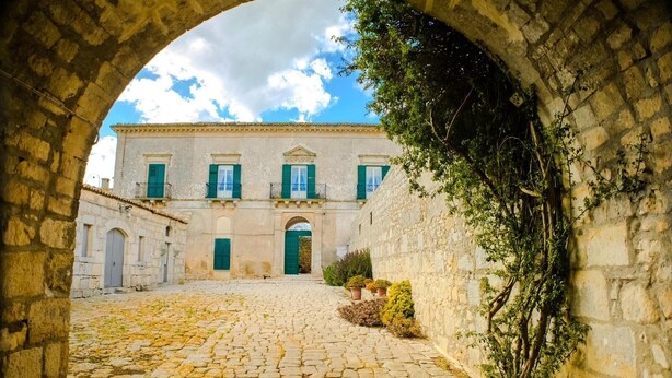 Un patio de piedra con un edificio al fondo, un arco y plantas en macetas.