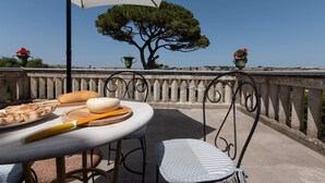 Un área de comedor al aire libre con una mesa preparada para dos personas, un paraguas grande y vistas a un árbol y la costa.