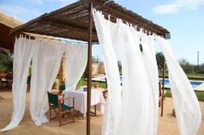 Uma área de estar coberta ao ar livre, com cortinas brancas, uma mesa e cadeiras.