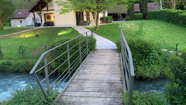 A wooden bridge over a river, leading to a building with a sloped roof and a paved pathway.