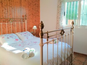 A bedroom with a wooden headboard, a bed with floral bedding, two rolled towels, and a window with curtains. - Villa Las Encinas on the Costa del Sol.        (Torrox)