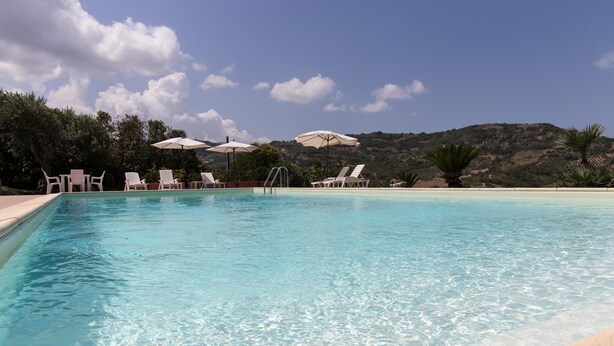 A clear blue pool with white lounge chairs and umbrellas, surrounded by greenery and a mountainous backdrop. - Wonderful villa with pool in a panoramic position at the gateway to Cilento. (Ogliastro Cilento)