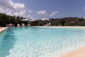 A clear blue pool with white lounge chairs and umbrellas, surrounded by greenery and a mountainous backdrop. - Wonderful villa with pool in a panoramic position at the gateway to Cilento. (Ogliastro Cilento)