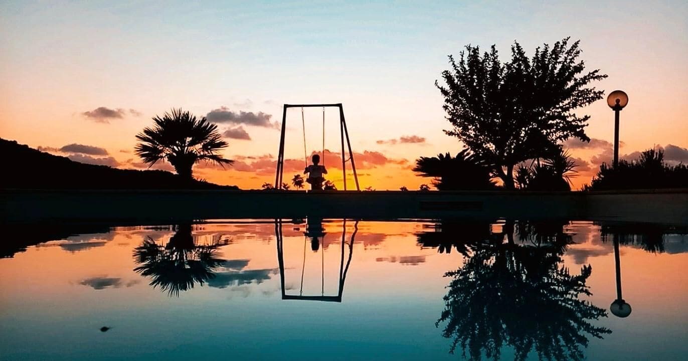 Une scène paisible en plein air, avec une balançoire, un lampadaire et un grand arbre qui se reflète dans une étendue d'eau calme, au coucher du soleil.