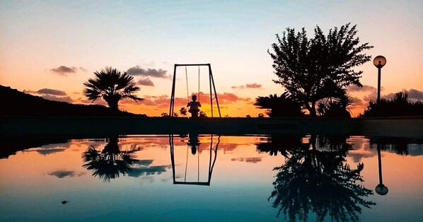 A tranquil outdoor scene with a swing set, a lamp post, and a large tree reflecting in a calm body of water during sunset.