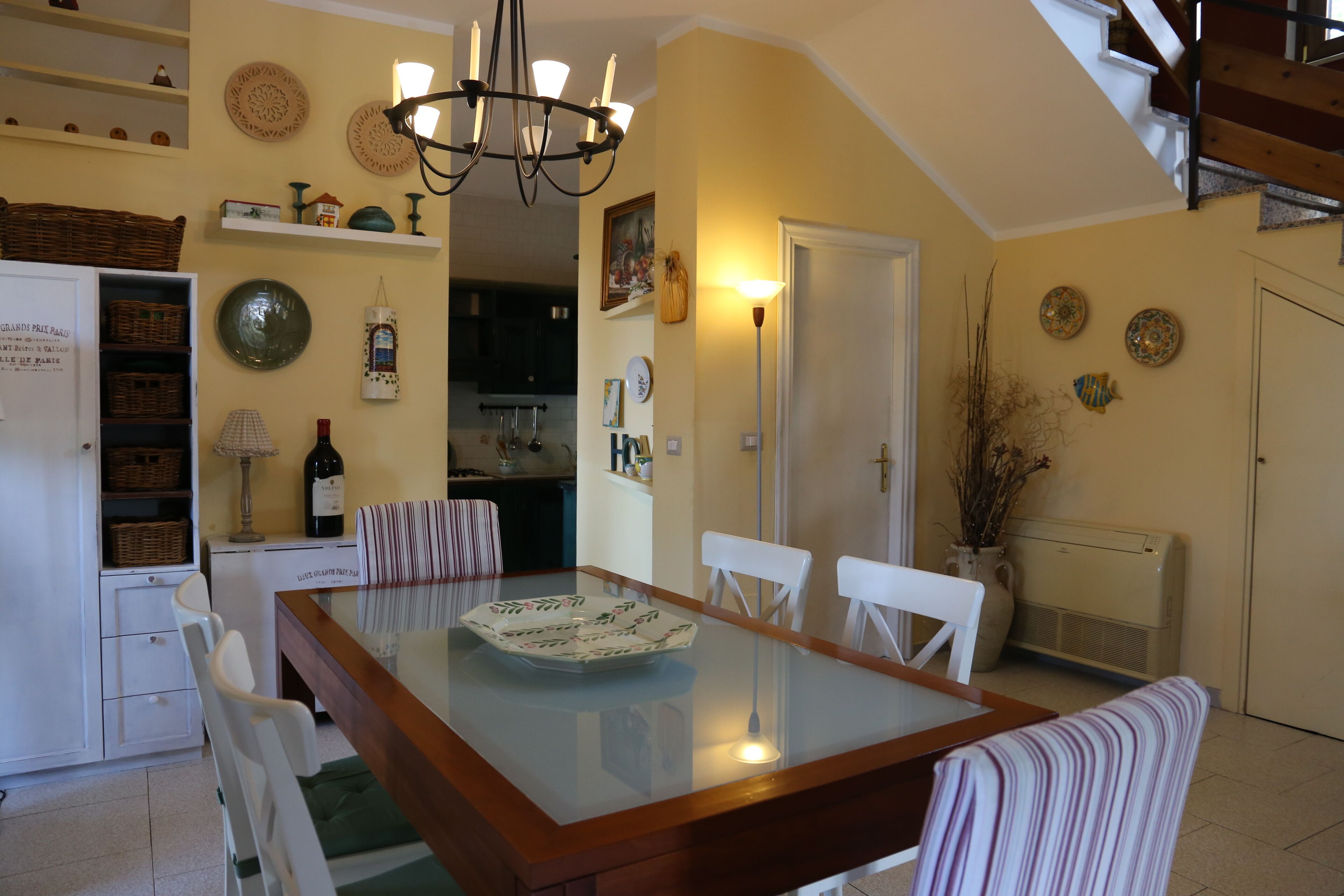 A dining area with a glass-top table, white chairs, a chandelier, and a staircase leading to an upper level.