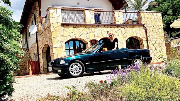 Une maison en pierre avec un balcon, une voiture garée devant et un jardin avec différentes espèces de plantes.